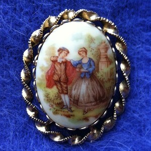 K&ouml;nnte beinhalten: Eine ovale Brosche in Goldt&ouml;nen mit einem wei&szlig;en Porzellankammeo, das eine gemalte Szene von zwei Personen in einem Garten zeigt. Das Kameo ist mit "Fragonard" signiert.
