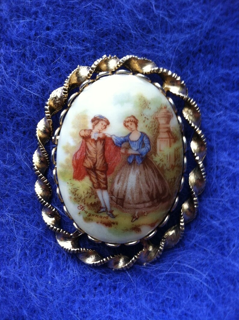 K&ouml;nnte beinhalten: Eine ovale Brosche in Goldt&ouml;nen mit einem wei&szlig;en Porzellankammeo, das eine gemalte Szene von zwei Personen in einem Garten zeigt. Das Kameo ist mit "Fragonard" signiert.