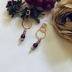 May include: A pair of gold hoop earrings with brown pearl accents. The earrings are hanging from a white surface with a flower and greenery in the background.