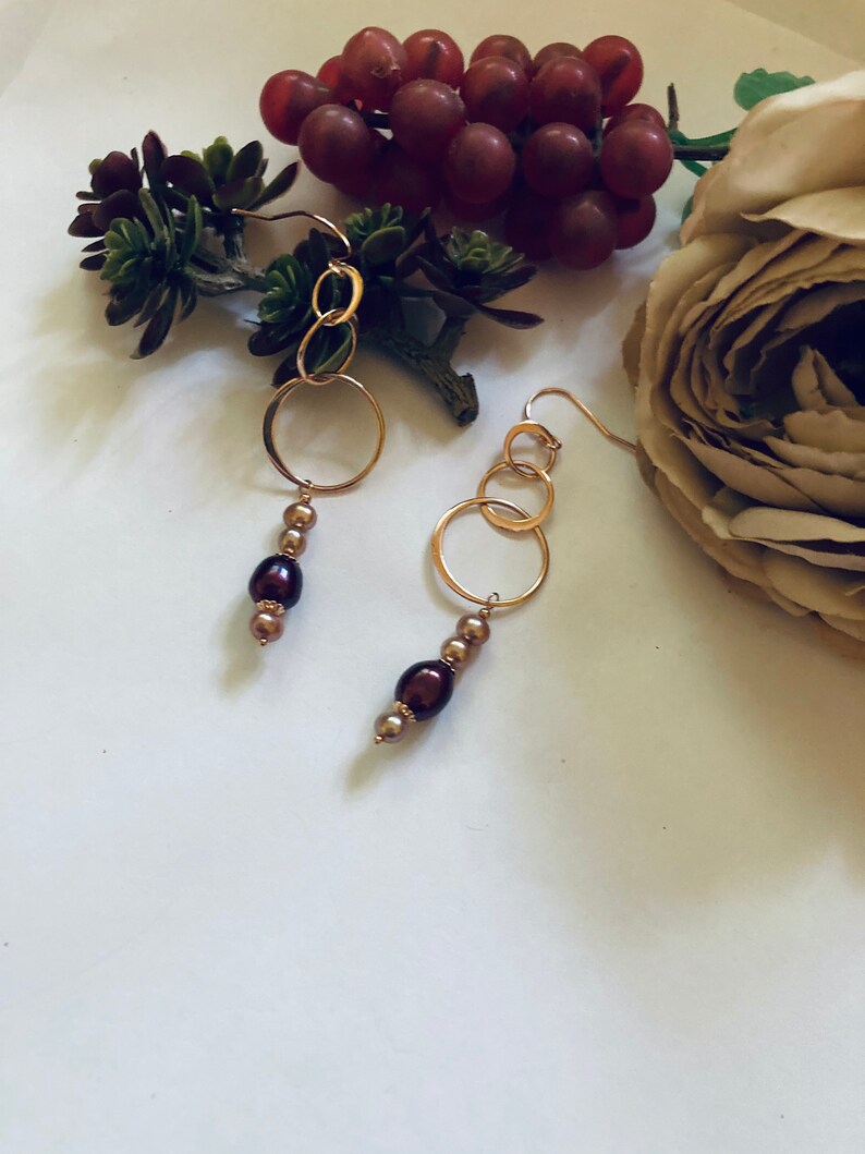 May include: A pair of gold hoop earrings with brown pearl accents. The earrings are hanging from a white surface with a flower and greenery in the background.