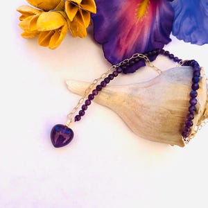 May include: A delicate gold chain necklace with a heart-shaped amethyst pendant. The necklace is made with small purple beads and is displayed on a seashell with yellow flowers in the background.