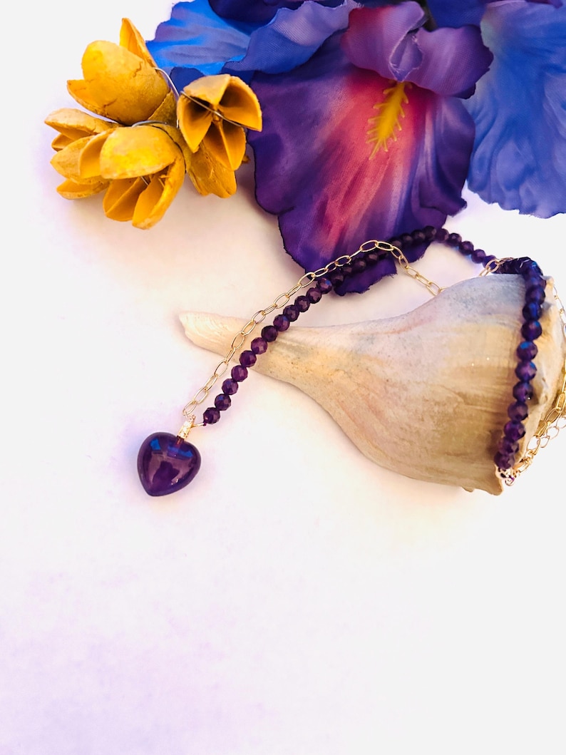 May include: A delicate gold chain necklace with a heart-shaped amethyst pendant. The necklace is made with small purple beads and is displayed on a seashell with yellow flowers in the background.