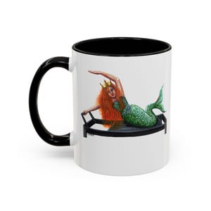 May include: A white ceramic mug with a black handle and interior rim. The exterior features a colorful illustration of a mermaid with long red hair and a gold crown, lying on a black Pilates reformer machine. The text C2023 is visible at the base.