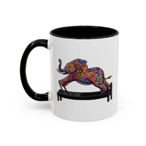 May include: A white ceramic mug with a black handle and interior rim. The exterior features a colorful, mosaic-style illustration of an elephant exercising on a black Pilates reformer machine. The elephant is depicted in vibrant shades of pink, blue, and yellow.