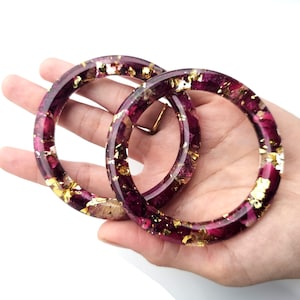 Pode incluir: Duas pulseiras redondas de resina transparente com um design floral vermelho escuro e folhas de ouro. As pulseiras são circulares e parecem ser feitas à mão. A resina é transparente, permitindo que os detalhes florais e as folhas de ouro sejam visíveis.