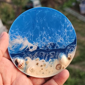 Puede incluir: Posavasos redondo de resina con una escena de playa. El posavasos presenta un diseño de olas azules y blancas, una playa arenosa y conchas marinas. El posavasos tiene aproximadamente 10 cm de diámetro.