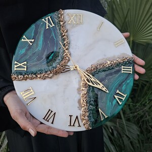 Può includere: Orologio da parete rotondo con un design marmorizzato bianco e verde acqua, accentuato da numeri romani dorati ed elementi decorativi dorati. Le lancette dell'orologio sono anch'esse dorate. L'orologio è tenuto davanti a uno sfondo verde.