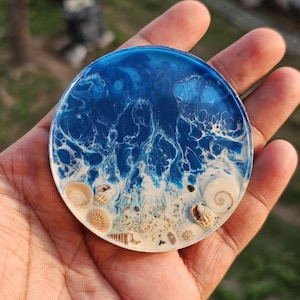 Puede incluir: Posavasos redondo de resina con un diseño de olas oceánicas azules y blancas. El posavasos presenta conchas marinas y arena incrustadas, creando una escena de playa en miniatura. El posavasos tiene aproximadamente 10 cm de diámetro.