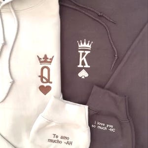 Könnte beinhalten: Zwei Kapuzenpullover, einer cremefarben und einer braun, mit Kronen- und Kartensymbolen. Der cremefarbene Kapuzenpullover hat ein Herz-Königin-Design, und der braune hat ein Pik-König-Design. Am Ärmel des cremefarbenen Kapuzenpullovers steht "Te amo mucho -AH", und am braunen Ärmel steht "I love you so much -HC".
