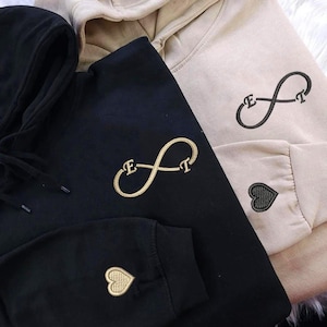 Puede incluir: Tres sudaderas con capucha en negro y beige. Cada sudadera presenta un símbolo de infinito bordado con iniciales y un corazón en la manga. La sudadera negra tiene bordados dorados, mientras que la sudadera beige tiene bordados negros.