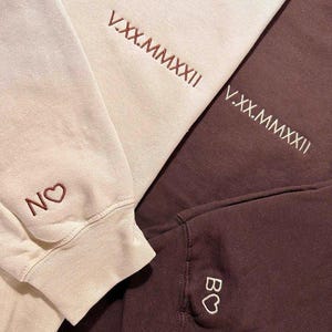 Könnte beinhalten: Nahaufnahme von drei Sweatshirts in Creme, Weiß und Braun. Jedes Sweatshirt hat gestickten Text und ein Herzsymbol. Das cremefarbene Sweatshirt hat "NO" und ein Herz. Die weißen und braunen Sweatshirts haben "V.XX.MMXXII" gestickt.