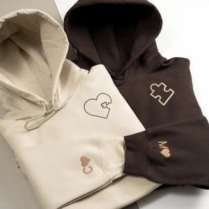 Könnte beinhalten: Zwei Kapuzenpullover, einer beige und einer braun, werden gezeigt. Der beige Kapuzenpullover hat ein Herz-Puzzleteil-Design auf der Vorderseite und ein Herz mit dem Buchstaben "C" am Bündchen. Der braune Kapuzenpullover hat ein Puzzleteil-Design und ein Herz mit dem Buchstaben "M" am Bündchen.