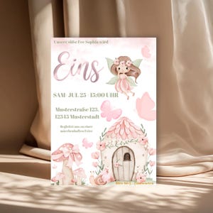 Op de afbeelding: Een roze en witte uitnodigingskaart met een fee&euml;nthema. De kaart bevat een fee&euml;nillustratie, vlinders en een paddenstoelenhuis. Tekst bevat de woorden "Eins" en evenementdetails.