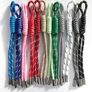 May include: A collection of twelve nylon cord zipper pulls in various colors including blue, pink, red, beige, light green, dark green, white, gray, and black. Each pull features a braided texture with reflective white threading and metal tips.
