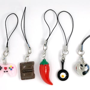 May include: Five colorful phone charms with black cords. The charms include a pink video game controller, a brown chocolate bar, a red chili pepper, a black frying pan with a white egg, and a silver heart.