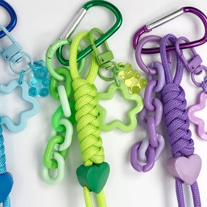 May include: Close-up of three colorful keychains with carabiner clips. Each keychain features a braided cord in blue, green, and purple, with matching heart-shaped charms. Additional charms include star and chain links in various shades of blue, green, and purple.