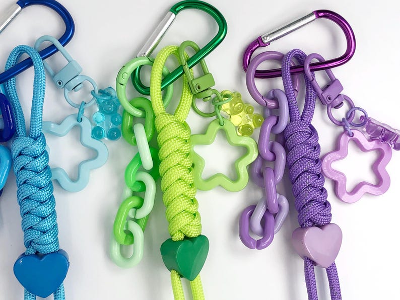 May include: Close-up of three colorful keychains with carabiner clips. Each keychain features a braided cord in blue, green, and purple, with matching heart-shaped charms. Additional charms include star and chain links in various shades of blue, green, and purple.