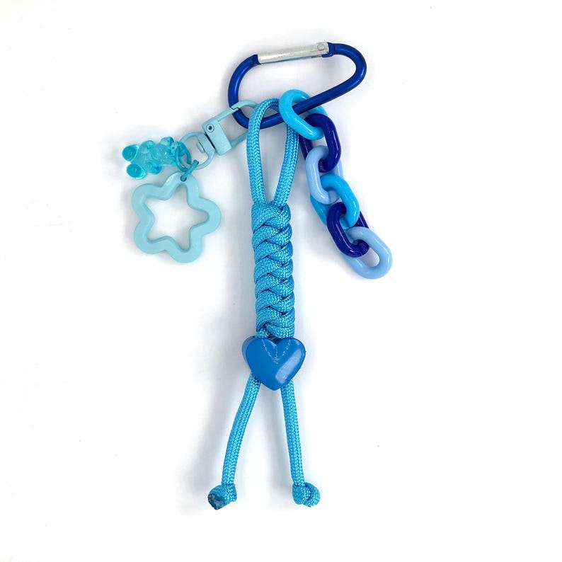 May include: A blue keychain with a carabiner clip, braided cord, and various charms. The charms include a star, a gummy bear, and a heart. The keychain features shades of blue and is designed for attaching to keys or bags.