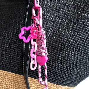 May include: A pink and white braided keychain with a pink carabiner clip. The keychain features a pink star, a chain of pink links, a pink heart, and a cluster of pink beads. The background is a black and tan woven bag.
