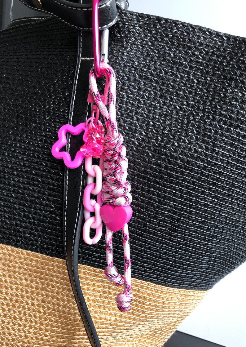 May include: A pink and white braided keychain with a pink carabiner clip. The keychain features a pink star, a chain of pink links, a pink heart, and a cluster of pink beads. The background is a black and tan woven bag.
