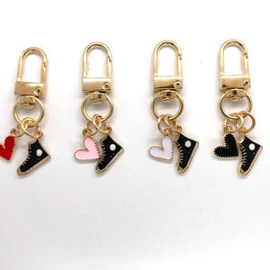 May include: Four gold-colored keychains with a lobster clasp. Each keychain has a charm with a black sneaker and a heart in red, pink, white, or black. The sneakers have white laces and a white circle.