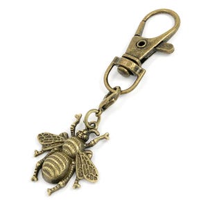 May include: Antique brass-colored bee charm with a lobster clasp. The bee has detailed wings and body stripes. The clasp is a swivel type, allowing for easy attachment to a keychain or bag.