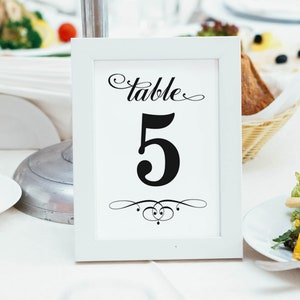May include: A white framed table number card with the word "table" in cursive script above the number "5". The number is in a bold, black font. A decorative flourish is below the number. The card is on a white tablecloth.