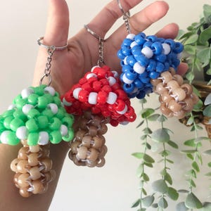 May include: Three beaded mushroom keychains with different colored tops. The tops are red, blue, and green. The stems are all a light brown color.