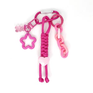 May include: A pink keychain with a variety of charms. The keychain includes a carabiner clip, a star-shaped charm, a heart-shaped charm, and a chain link charm. The keychain is made of metal and plastic.