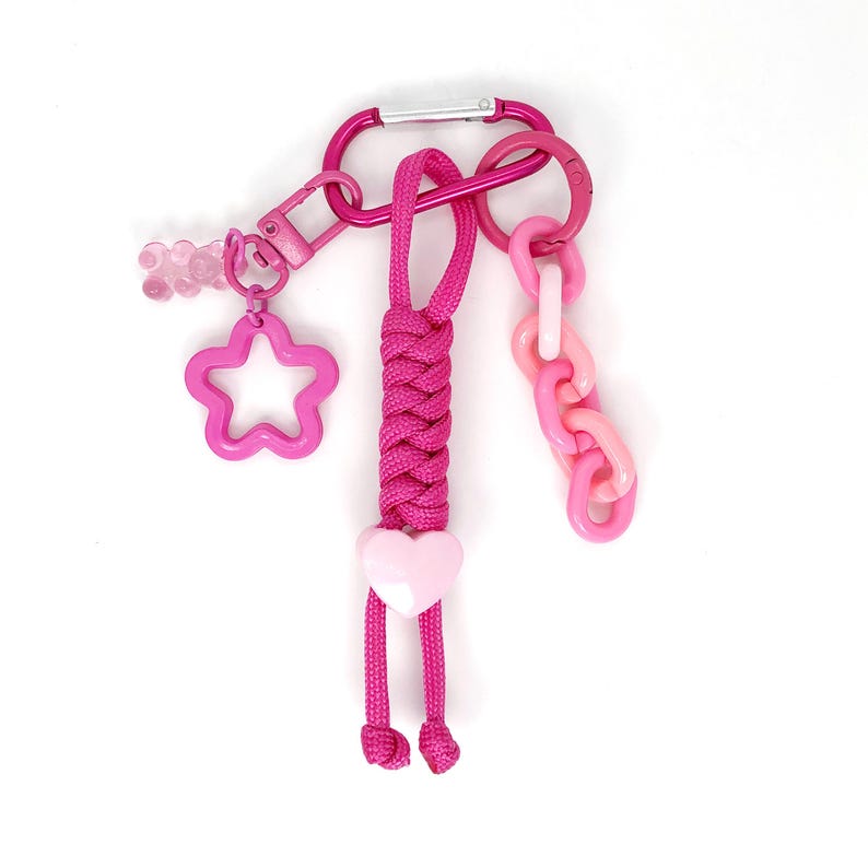 May include: A pink keychain with a variety of charms. The keychain includes a carabiner clip, a star-shaped charm, a heart-shaped charm, and a chain link charm. The keychain is made of metal and plastic.