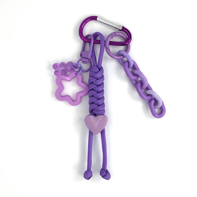 May include: A purple keychain with a braided cord, heart charm, star-shaped charm, and a chain. The keychain has a purple carabiner clip and a small clasp. The charms are various shades of purple.