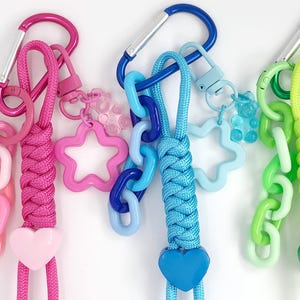 May include: Three colorful keychains with carabiner clips. One is pink, with a pink chain, star, and heart charm. Another is blue, with a blue chain, star, and bear charm. The last is green, with a green chain, star, and heart charm.