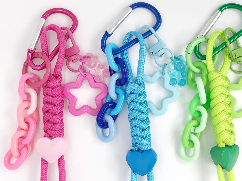 May include: Three colorful keychains with carabiner clips. One is pink, with a pink chain, star, and heart charm. Another is blue, with a blue chain, star, and bear charm. The last is green, with a green chain, star, and heart charm.