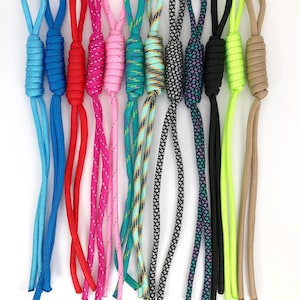 May include: A collection of twelve paracord zipper pulls in various colors and patterns, including solid blue, red, pink, black, neon green, and tan, as well as multi-colored braided designs, each featuring a looped top and a wrapped knot.