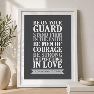 May include: A framed print with a white border displays the text "BE ON YOUR GUARD, STAND FIRM IN THE FAITH, BE MEN OF COURAGE, BE STRONG, DO EVERYTHING IN LOVE" in white and gray lettering on a dark gray background. The scripture reference "1 Corinthians 16:13-14" is at the bottom.