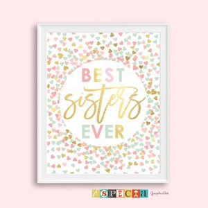 May include: A framed print with the words "BEST SISTERS EVER" in gold script, surrounded by a confetti of pastel hearts. The background is a light pink, and the frame is white. The print is a decorative piece.