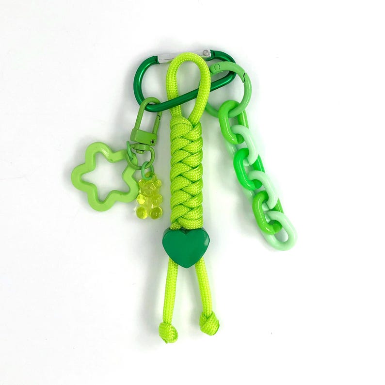 May include: A green keychain with a braided neon green cord, a green heart charm, and a green carabiner clip. It features a light green flower-shaped charm, a small gummy bear charm, and a chain of green and white links.