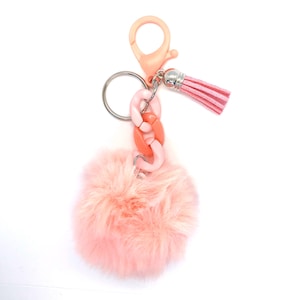 May include: A pink keychain with a fluffy pom-pom, a chain link, and a tassel. The keychain has a lobster clasp and a silver keyring. The pom-pom is made of soft, faux fur. The chain link is a mix of pink and coral colors.