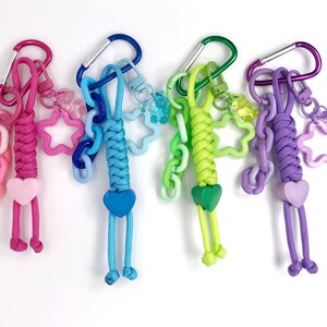 May include: Four colorful keychains with carabiner clips. Each keychain features a braided cord body, a heart-shaped charm, and dangling cords. The keychains are in pink, blue, green, and purple, with star and chain accents.