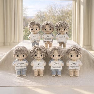May include: Seven crocheted dolls with distinct hairstyles, each holding a sign with a name and quote, such as "RM Speak yourself". The dolls are dressed in light blue and beige outfits. The dolls are displayed on a white surface.