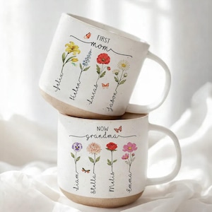 Puede incluir: Dos tazas de cerámica blanca apiladas. La taza superior dice "First Mom" y tiene diseños florales con nombres. La taza inferior dice "Now Grandma" y tiene diseños florales con nombres. Las tazas tienen una base beige y un asa blanca.