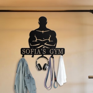 May include: Black metal wall-mounted gym organizer with a silhouette of a muscular figure and the text "SOFIA'S GYM". It features hooks holding a denim jacket, headphones, jump rope, and a white towel. The gym setting includes exercise equipment.