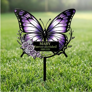 Könnte beinhalten: Ein schwarzer Metall-Gartenstecker mit einem großen, violett-weißen Schmetterling mit detaillierten Flügelmustern. Eine rechteckige Plakette in der Mitte zeigt den Text MARY, Forever cherished, sowie die Jahreszahlen 1938-2026.