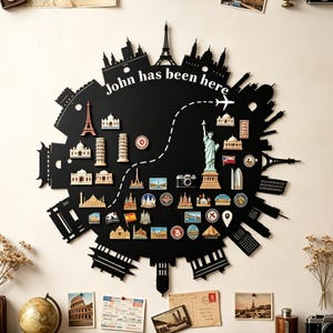 Puede incluir: Un expositor de pared circular negro con una silueta de horizonte urbano y el texto John has been here. La superficie está cubierta con varios imanes pequeños de temática viajera, incluyendo monumentos como la Torre Eiffel y la Estatua de la Libertad.