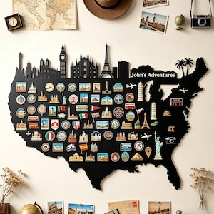 Può includere: Una mappa da parete nera degli Stati Uniti con una silhouette dello skyline nella parte superiore e il testo "John's Adventures". La mappa è ricoperta da numerosi magneti souvenir colorati, tra cui monumenti, bandiere e piccoli aerei.