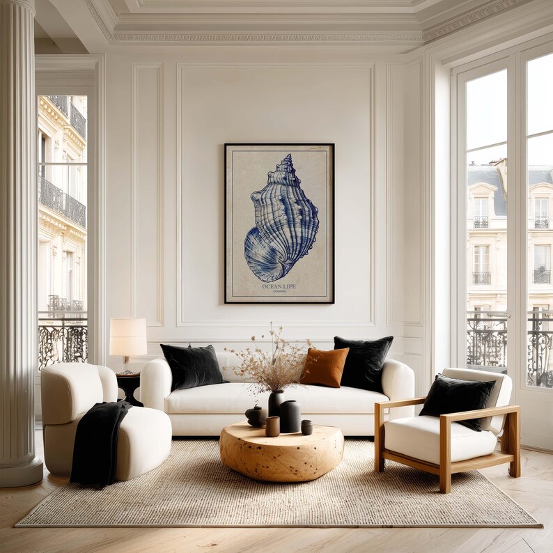 Large nautical shell art print in blue on textured beige paper, framed above a sofa in a bright living room with tall windows and a balcony view.