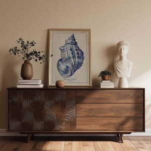 Mid-century styled seashell art print in blue on textured paper resting on a sideboard with sculptural decor and stacked books.