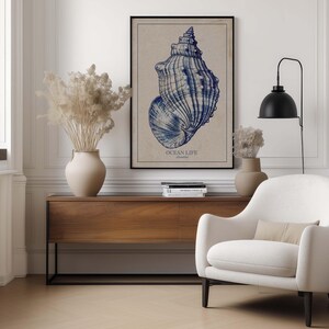 Elegant seashell wall art print in indigo and beige above a wooden console table with neutral vases and a lamp next to a cozy armchair.