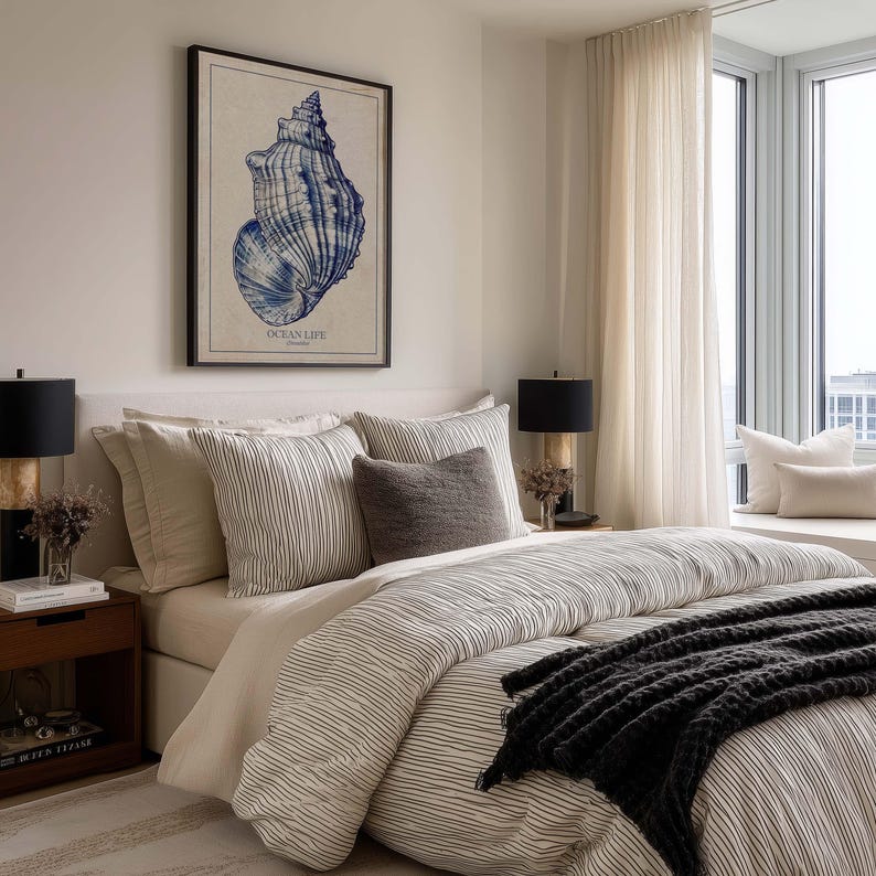 Framed blue shell art print on warm beige paper hanging above a neatly made bed in a modern neutral bedroom.
