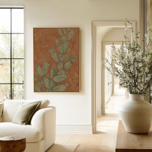 Botanical prickly pear cactus in green and warm brown, rustic vintage mood, presented as a framed poster in an airy living room near an archway.